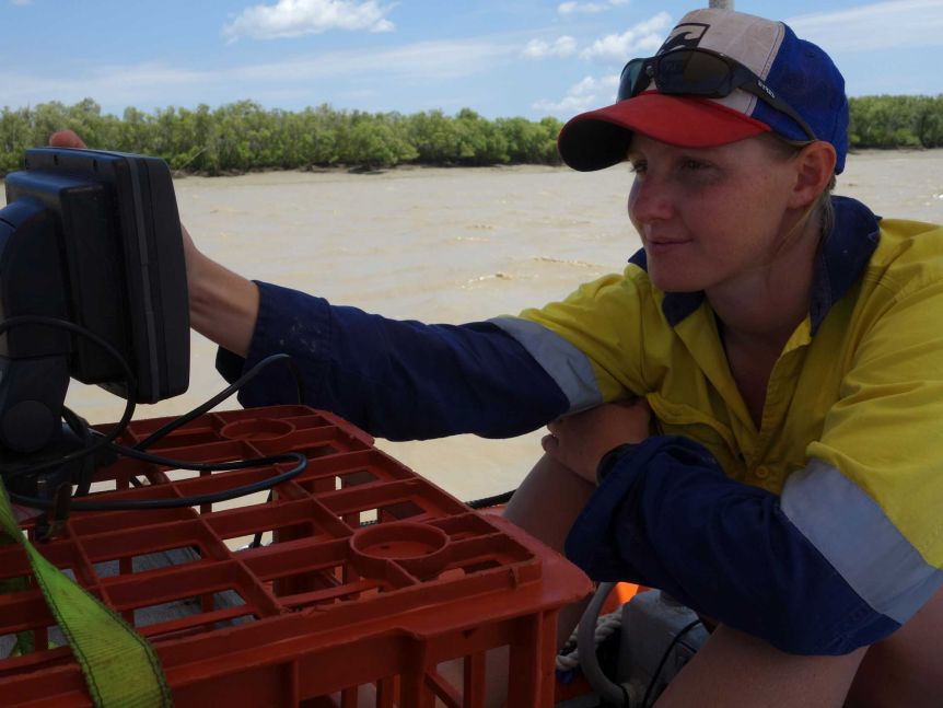 A young woman wearing a cap and high-vis gear looks at a small monitor that is perched on a milk crate.