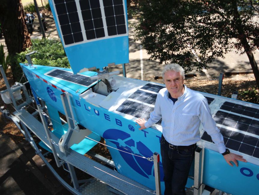 A silver-haired man in business attire leans against a bright blue boat covered in solar panels.