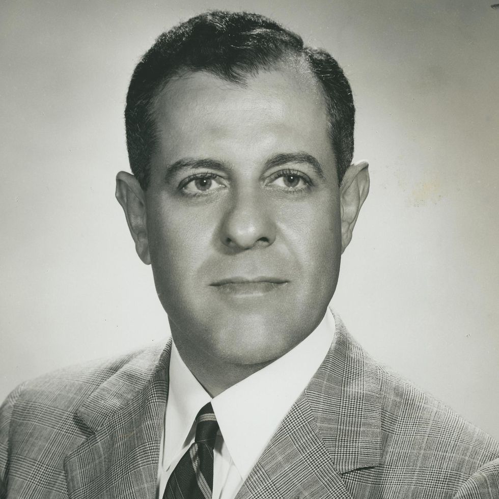 A man with dark, closely cropped hair and wearing a glen plaid suit jacket smiles bemusedly.