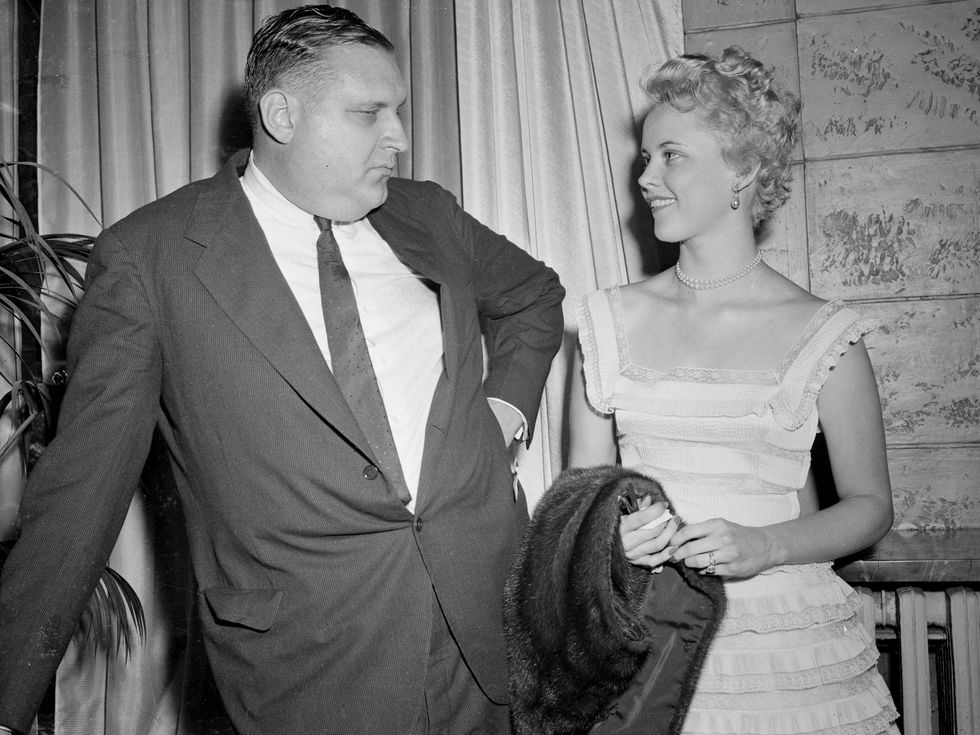 A man in a suit stands next to a woman in a white sleeveless dress holding a fur coat.