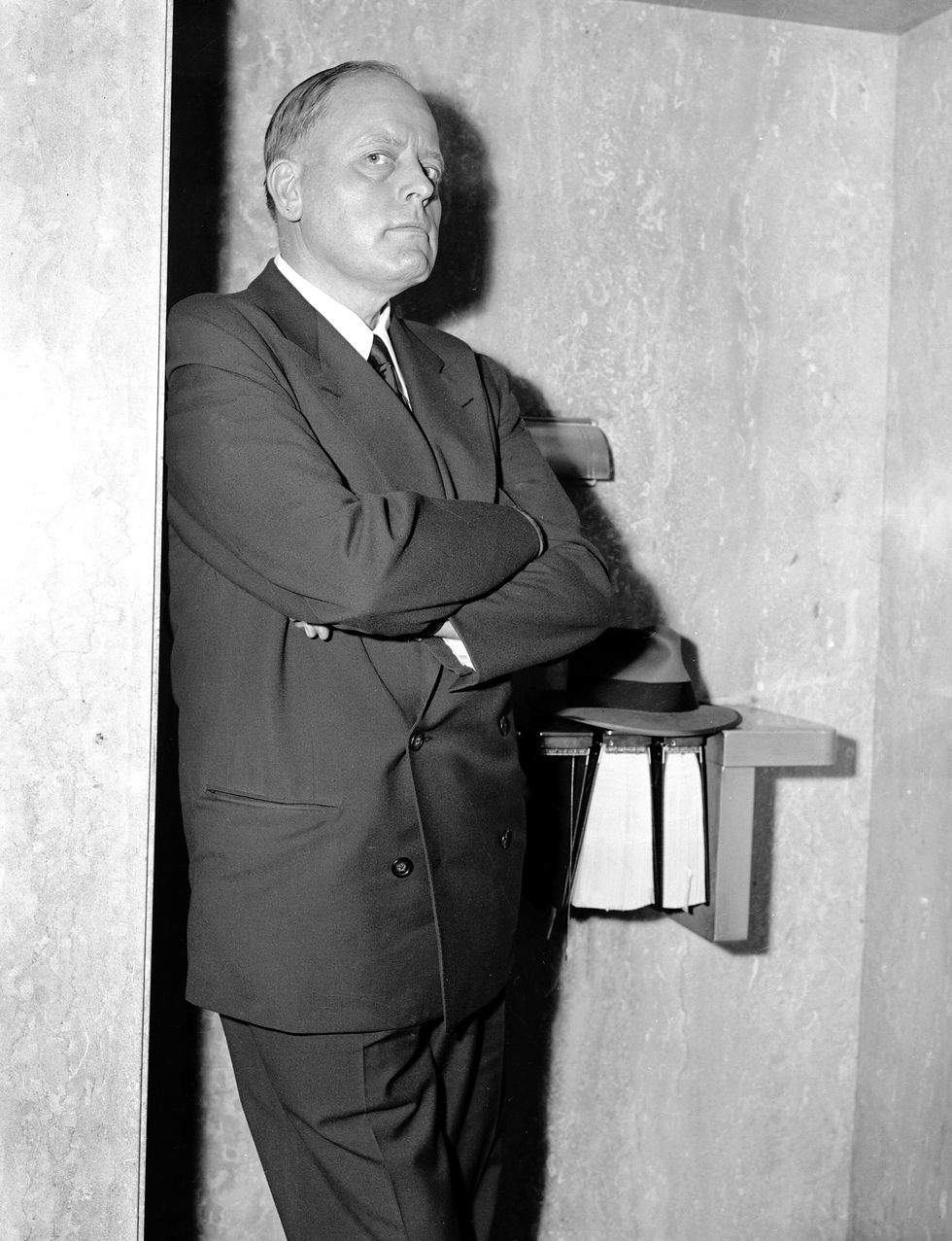A man in a double-breasted suit, arms folded over his chest, and with a haughty expression on his face, leans against a wall in a telephone kiosk.