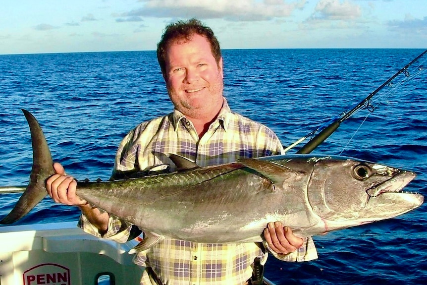 A man stands on a boat that is at sea. He is holding a large fish in his hands. He is smiling.