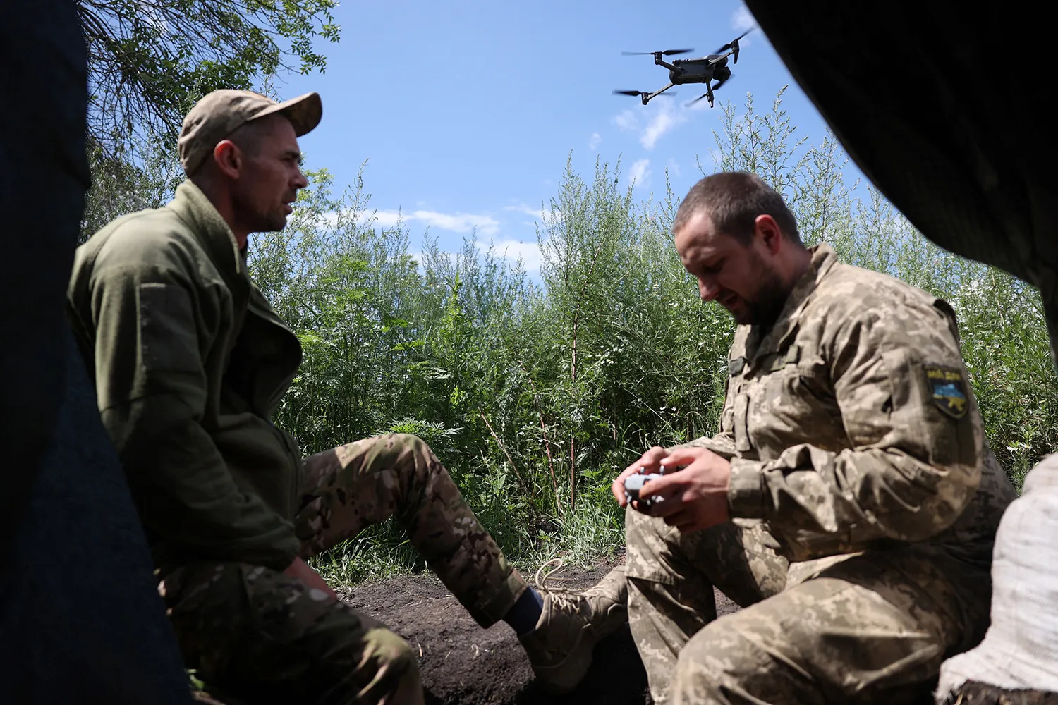 Two servicemen are pictured on the frontlines with a drone in the background.